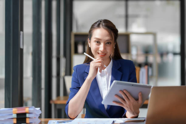 istock Asian business woman sitting happily with laptop and using tablet and taking intense notes on the work clipboard and smiling happily on the assignment. 1829694925