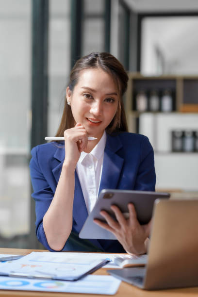 istock Asian business woman sitting happily with laptop and using tablet and taking intense notes on the work clipboard and smiling happily on the assignment. 1829694937