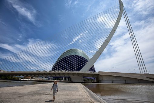 Assut de l'Or Bridge is a dazzling white single-pylon cable-stayed bridge, located in the City of Arts and Sciences complex, Valencia, Spain. A woman walking by. L'Àgora in the background.