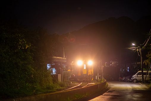 A yellow diesel train passes through the village at night with its lights on. Along the Pingxi line, there are river valleys, potholes and waterfalls. Taiwan