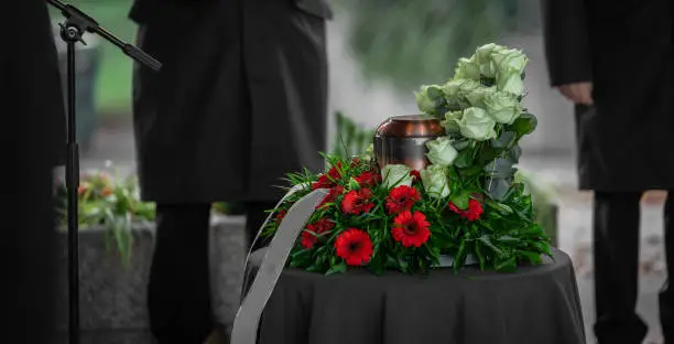 Metal urn or funeral container with ash of a deceased person at a memorial service. Undertakers seen in the back and a beautiful bouquet surrounding the urn.