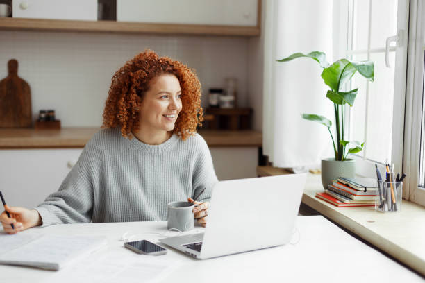 istock Young inspired redhead screenwriter or author working at kitchen table with laptop, smartphone and coffee cup, looking aside through window inventing new plot and dialogues, making notes in copybook 1829867977