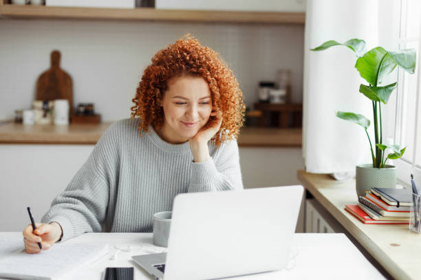 istock Cute girl with red curls watching online tutorial or lesson on laptop, smiling listening and looking attentively, making notes in copybook, sitting at kitchen table with smartphone and cup of coffee 1829867978
