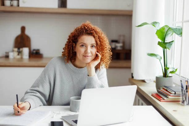 istock Young ginger headed owner of online store working from home at kitchen table, using laptop and wireless connection, looking at camera, writing in copybook, making business plan for new project 1829867982