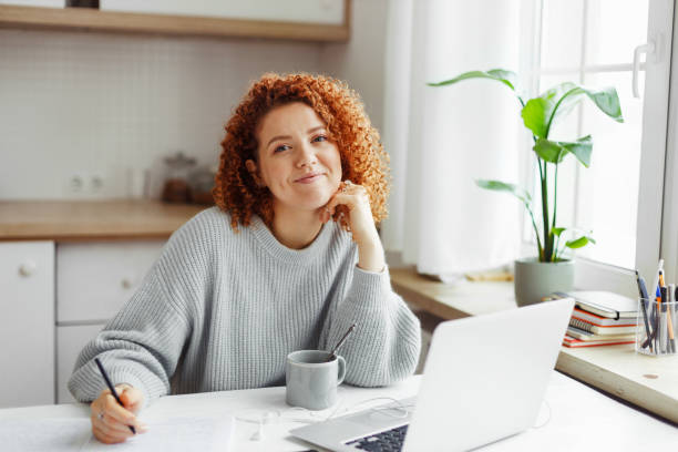 istock Cute curly redhead PR-manager of online store creating content plan working online at home, sitting at kitchen table with laptop and coffee cup, writing in copybook, making notes, looking at camera 1829868502