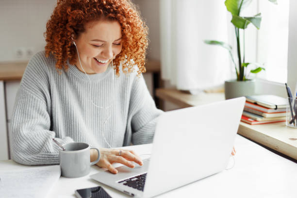 istock Student with red curls studying at home, having rest video-chatting with her mates on laptop, sitting at kitchen table with coffee cup and smartphone in wired earphones, laughing looking at screen 1829869009