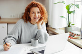 Portrait of smiling redhead curly female student studying at home, doing home work in front of laptop, preparing for exams, sitting at kitchen table with pen in hands, looking at camera