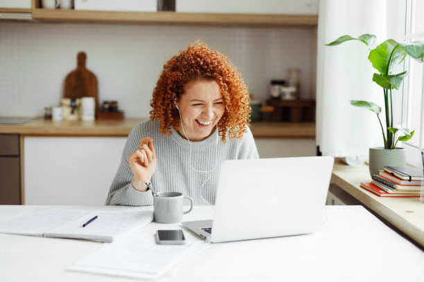 istock Funny laughing redhead female tutor having video chat with her student, sitting at kitchen table in front of laptop with copy book, coffee cup and smartphone on desk, explaining new topic 1829870558