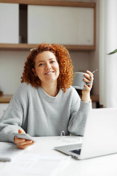 Happy smiling woman having coffee break holding cup in hand and using smartphone, checking social media updates, shopping, browsing web pages sitting in front of opened laptop at kitchen table
