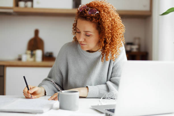 istock Redhead female interpreter freelancer with curly hair working from home, writing translation in her copybook sitting at kitchen table in front of laptop, translating article for web site 1829873508