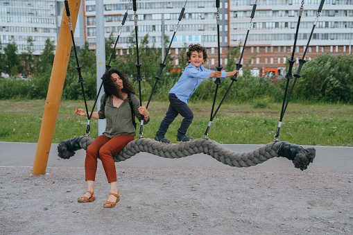 Active curly little boy riding on swing at playground with mom. Childhood, family leisure. Happy children.Young babysitter entertains little kid outdoors.