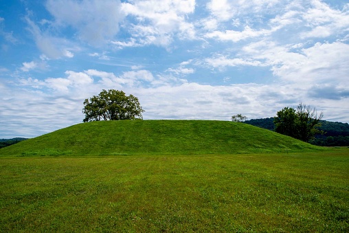Grass covered prehistoric native American Hopewell culture burial mound at Seip Earthworks in Ohio under a dramatic cloudy sky with neatly cut green grass foreground and tree. No people, with copy space.