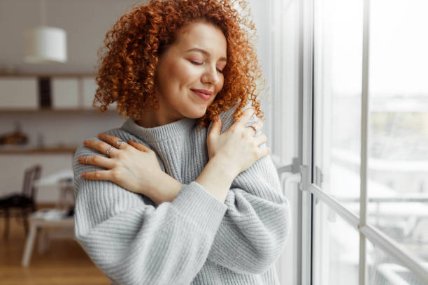 horizontal image of pretty redhead female with closed eyes wearing rings on fingers, hugging herself, touching new soft sweater, enjoying comfort of fabric, standing next to panoramic window at home - självförtroende bildbanksfoton och bilder