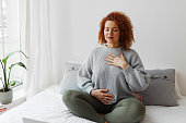 Portrait of plus-size curly redhead female in casual clothes doing pranayama technique sitting on bed next to window with closed eyes, putting hands on chest, breathing slowly and deeply