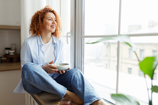 Young woman with red curly hair in jeans and shirt sitting on window sill looking through big window on city holding bowl of breakfast in hands, smiling noticing something funny outside
