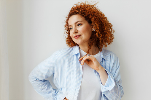 Pensive smiling woman with sad eyes and red curly hair in blue casual shirt posing against white wall at home, looking aside, recollecting bitter sweet memories of her ex-boyfriend, playing with hair