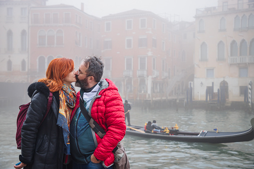 Beautiful middle aged heterosexual married couple in warm clothing standing embraced by Grand Canal and kissing, foggy day in winter, visiting Venice in Italy together