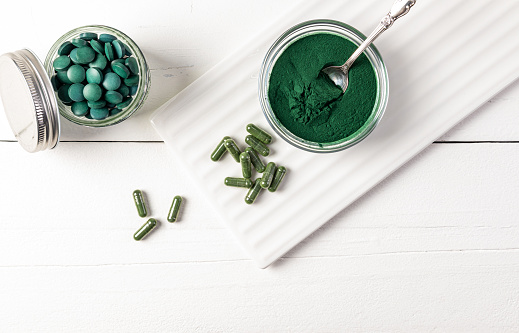 Spirulina powder, tablets, capsules in various jars on white wooden background and ceramic stand. Top view. Healthy nutritional supplements