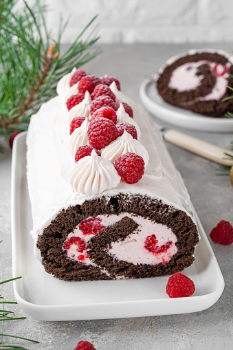 Chocolate christmas roll cake with cream and fresh raspberries on a white plate on a gray background. Christmas yule log cake