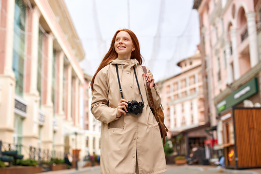 Young smiling woman tourist walks around city in spring, holds retro camera. Redhead female on trip, in casual urban style outfit. Portrait of caucasian lady outdoors, alone