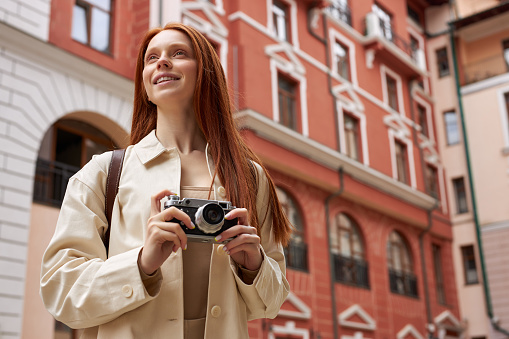 Redhead photographer woman with retro camera in hands looking at urban old architecture, taking pictures, discover new places, walking alone in city. Adult female with long natural red hair enjoy trip