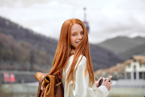 good-looking red-haired woman with retro camera in hands on trip, in mountains, in casual coat or jacket. traveling alone, spend holidays, take photos.