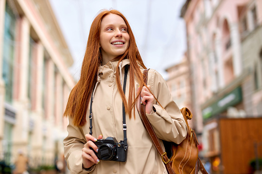 Relaxed redhead woman traveler with warm smile take photo on film retro camera while walking in urban town, portrait of trendy friendly lady in casual wear with backpack at spring cold weather