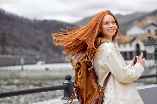 Young redhead woman smiling happy using vintage camera walking at mountains, female turned the head back and look at camera, in casual outfit. portrait of cute lady with leather travel backpack