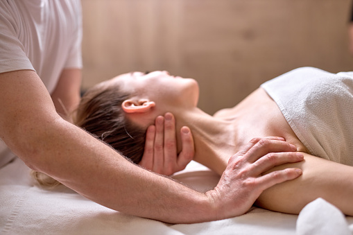 Professional cropped therapist doing healthy massage on neck and shoulders to female client lying on back, side view. at spa salon