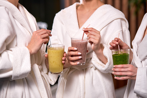 Cropped three women enjoy drinking healthy beverage during spa treatment wearing bathrobes, drinking freshly squeezed juice while spending weekend in wellness center.