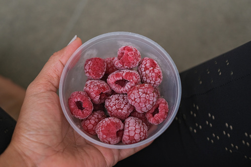 Close-up  POV shot unrecognizable person sitting on the ground, her hand holding a small bowl plastic container with frozen raspberry
