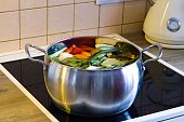 A stainless steel pot on an electric stove with a ceramic cooktop cooking organic chicken broth. The broth contains vegetables, chicken, bell pepper, celery, leeks, celery leaves, bay leaves, garlic, onion, and carrots.