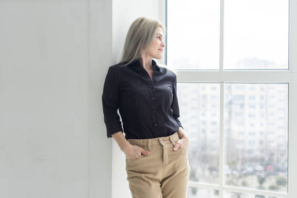 istock Young elegant woman thinking something while standing near office window outside, female employer while looking forward to meeting with international partners 1829985968