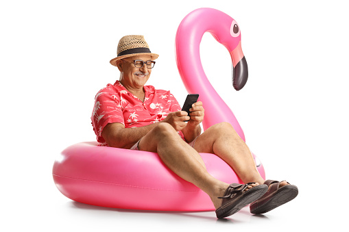 Mature male tourist sitting on a famingo swimming ring and using a smartphone isolated on white background