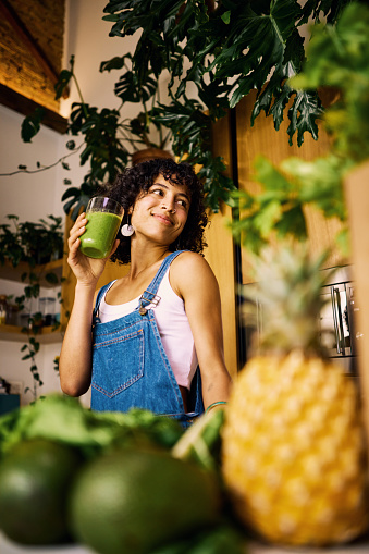 Happy Woman Enjoying A Fresh Green Smoothie In A Sunny Kitchen Decorated With Indoor Plants Promoting A Healthy Lifestyle And Sustainable Living