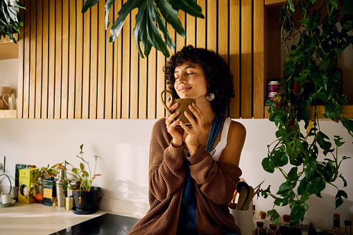 Content Woman Savoring The Aroma Of Her Morning Coffee In A Well-Lit Kitchen With Modern Wooden Accents And Lush Houseplants, Embracing A Moment Of Serenity, Symbolizing Mindfulness And The Joy Of Simple Pleasures In Everyday Life