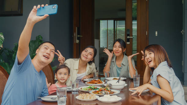 istock Young Asian family using mobile phone taking selfie and having fun sitting at dining table at backyard outside home. Multi-generation family enjoying spending together. 1830013387