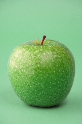 Stock photo showing close-up view of single, green Granny Smith apple with shiny, speckled skin against a pale green background.