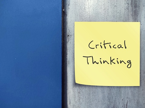 Blue wall with handwritten sticky note CRITICAL THINKING - Ability to think clearly rationally about what to do - analyzing facts to understand a problem or topic thoroughly