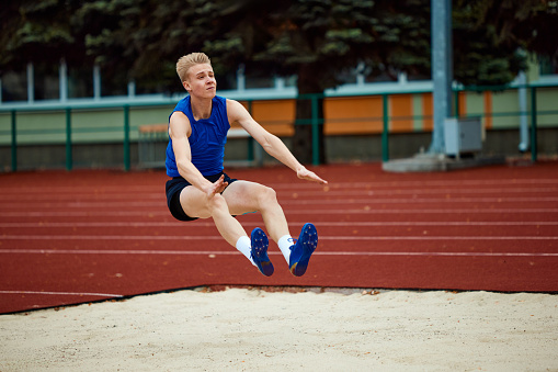Young male runner in sportswear running fast and jumping through hurdle while training at stadium track side view