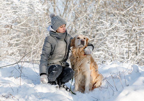 Teenage Girl And Golden Retriever Sit Together In Snow-Covered Forest During Winter