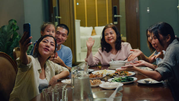 istock Asian family using mobile phone taking video call and having fun dinner sitting at dining table at backyard outside home at night. Multi-generation family enjoying spending together. 1830080405