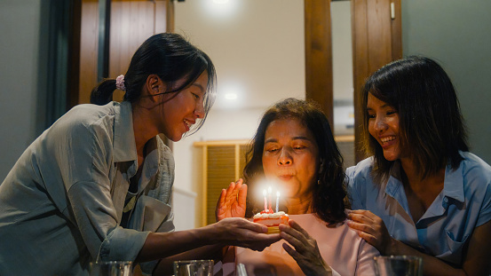 Asian family blowing Birthday candle on a cake while celebrating and having fun sitting at dining table at backyard outside home at night. Multi-generation family enjoying spending together concept.