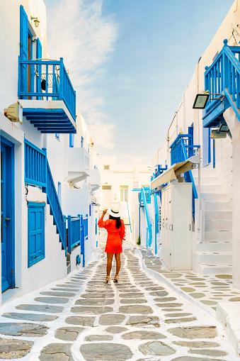 Young women in red dress walk on the street of Mykonos Greek village in Greece, colorful streets of Mikonos village. Asian woman visits Mykonos village during vacation holiday in Europe