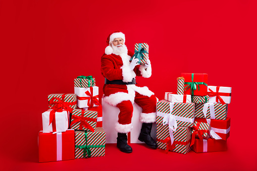Full length photo of cool funky cheerful man santa claus sit platform hand hold giftbox isolated on red color background year.