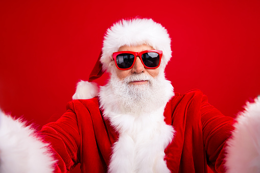 Photo of positive cheerful grandfather in santa costume in glasses making selfie on smartphone camera isolated on red color background.