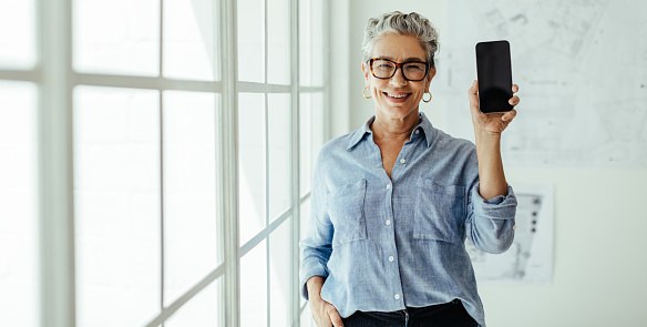 Happy female architect holding a mobile phone with a blank screen in her office. Senior design expert smiling at the camera as she recommends the use of smart technology in her profession.
