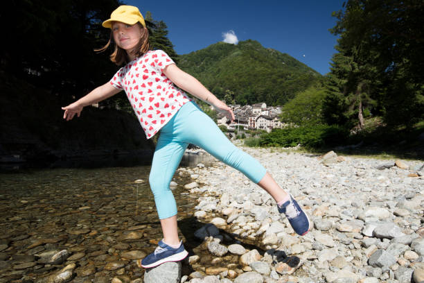 A young girl is playing along the creek called Cervo, in Rosazza. She 's balancing on one foot on a rock. It's summertime even in the mountains and the day's crystal clear