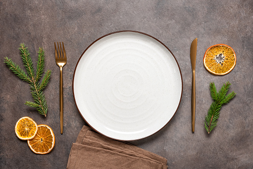 Christmas minimal table setting. An empty white plate, gold cutlery, a linen napkin, dry orange slices and spruce branches. Dark brown background. Top view, flat lay.