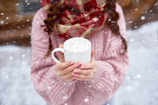 Young woman holding cup of hot chocolate with marshmallows outdoors in winter day